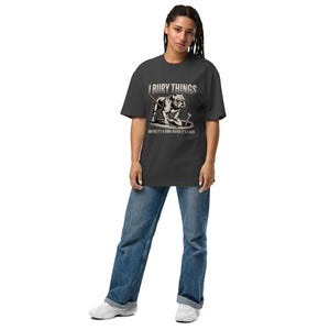 Op de afbeelding: Donkergrijs t-shirt met de tekst "I BURY THINGS" en een afbeelding van een bulldog die graaft. Daaronder staat "MAYBE IT'S A BONE MAYBE IT'S A BODY". Het shirt wordt gedragen met een blauwe jeans en witte sneakers.