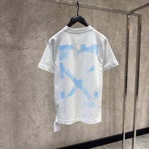 May include: White t-shirt with a light blue graphic design on the back. The design features a cross and rectangular shapes. The shirt is hanging on a black hanger against a neutral background.