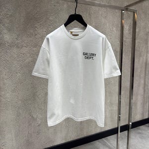Puede incluir: Una camiseta blanca de manga corta y cuello redondo, colgada de una percha negra. El texto "GALLERY DEPT." est&aacute; impreso en negro en la parte delantera. La camiseta se exhibe en una percha met&aacute;lica contra una pared texturizada.