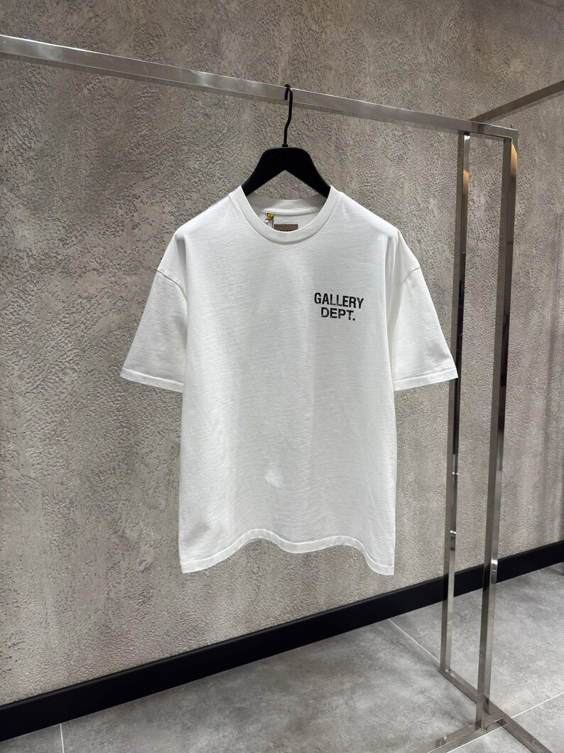 Puede incluir: Una camiseta blanca de manga corta y cuello redondo, colgada de una percha negra. El texto "GALLERY DEPT." est&aacute; impreso en negro en la parte delantera. La camiseta se exhibe en una percha met&aacute;lica contra una pared texturizada.