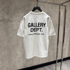Puede incluir: Una camiseta blanca con el texto "GALLERY DEPT. HOLLYWOOD, CA." impreso en negro. La camiseta est&aacute; colgada de una percha negra en una percha met&aacute;lica. El fondo es una pared gris texturizada.