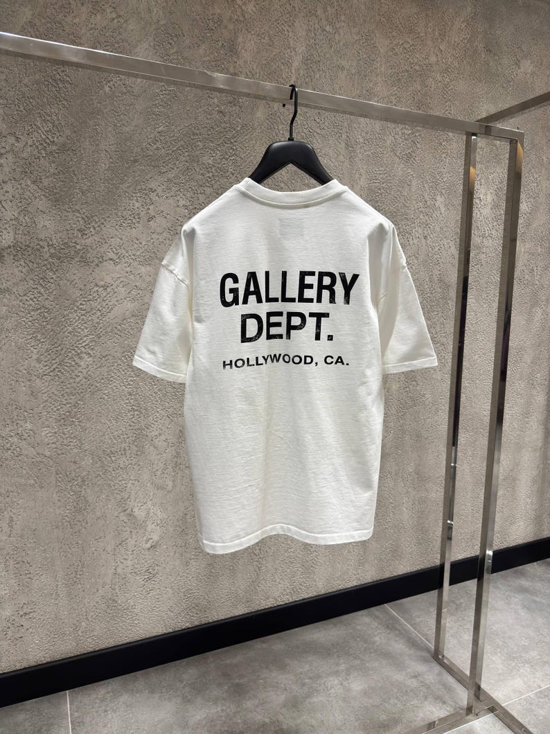 Puede incluir: Una camiseta blanca con el texto "GALLERY DEPT. HOLLYWOOD, CA." impreso en negro. La camiseta est&aacute; colgada de una percha negra en una percha met&aacute;lica. El fondo es una pared gris texturizada.