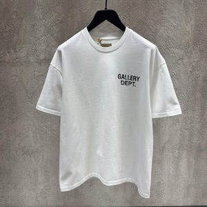 Puede incluir: Camiseta blanca de manga corta con el texto "GALLERY DEPT." impreso en negro en la parte delantera. La camiseta est&aacute; colgada de una percha negra sobre un fondo gris.