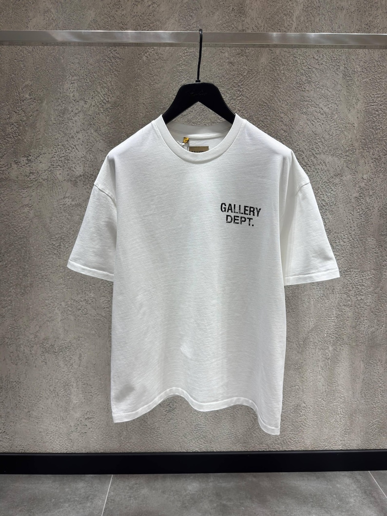 Puede incluir: Camiseta blanca de manga corta con el texto "GALLERY DEPT." impreso en negro en la parte delantera. La camiseta est&aacute; colgada de una percha negra sobre un fondo gris.