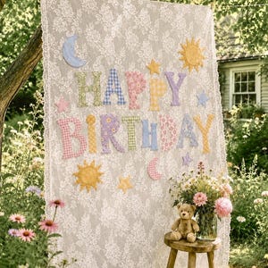May include: A rectangular white lace fabric backdrop featuring the text HAPPY BIRTHDAY in multicolored fabric letters. The design includes appliqued sun, moon, and star shapes in various patterns and colors, suspended outdoors.
