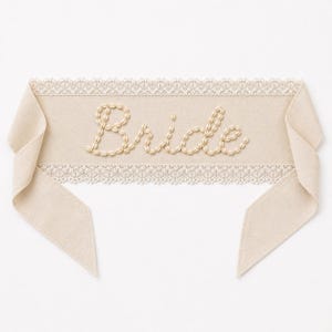 May include: A beige fabric sash with the word "Bride" embroidered in a cursive style. The sash has a delicate lace trim along the top and bottom edges. The ends of the sash are cut at an angle.