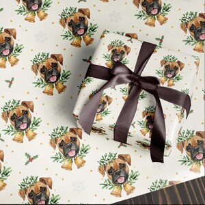 Puede incluir: Papel de regalo con un patrón repetido de un perro Boxer con una corona y campanas. El fondo es blanquecino con copos de nieve y puntos dorados. Un regalo envuelto con una cinta de raso marrón está en la imagen.