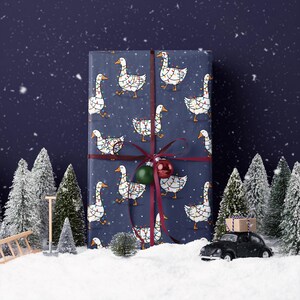May include: A wrapped gift with navy blue wrapping paper featuring white geese decorated with Christmas lights. The gift is tied with a burgundy ribbon and adorned with a green and red ornament. The scene is set in a snowy winter landscape.