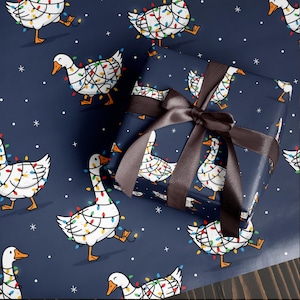 May include: Gift wrap featuring a repeating pattern of white geese adorned with colorful Christmas lights on a navy blue background. A wrapped gift with a brown satin ribbon is in the image. White snowflakes and stars are scattered across the background.