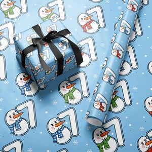 Pode incluir: Uma caixa de presente embrulhada com uma fita preta e um rolo de papel de embrulho. Ambos apresentam um padrão azul claro com bonecos de neve com cachecóis e o número 7. O fundo é azul claro com flocos de neve.