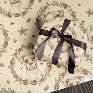 May include: Gift wrapped in festive paper featuring a repeating pattern of a Boxer dog, pine cones, stars, and greenery. The gift is tied with a dark brown satin ribbon and bow. The wrapping paper has a vintage, sepia-toned aesthetic.
