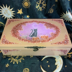 May include: A wooden jewelry box with a painted scene of a black cat sitting on pumpkins under a purple moon. The box is beige with a decorative metal clasp and feet. The lid is framed with autumn leaves.