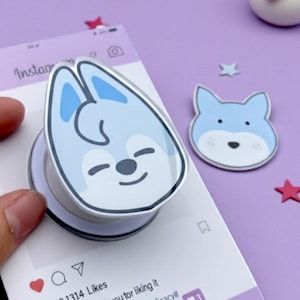 May include: A light blue and white pop-up phone grip with a cartoon fox face, held over a purple Instagram-themed card. The card has text and social media icons. Scattered around are star-shaped and donut-shaped decorations.