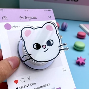 以下が含まれることがあります： 紫色のInstagramをテーマにしたカードに付いた、白い猫の形をした電話グリップ。カードには、ハート、コメント、共有アイコン、テキスト「Album. Thank you for liking it.」と「#Share daily life# #Today# #Delicacy#」が表示されています。