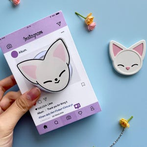 Może przedstawiać: Biały uchwyt do telefonu z motywem kota i pasujący biały magnes z motywem kota z różowymi akcentami. Uchwyt do telefonu jest przymocowany do fioletowej karty z motywem Instagrama. Karta zawiera tekst i jest trzymana w dłoni.