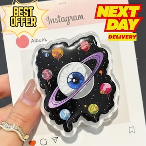 May include: A clear, plastic phone grip featuring a cosmic eye design. The eye has a blue iris and is surrounded by planets and a purple ring. The background is black with white stars. The words "BEST OFFER" and "NEXT DAY DELIVERY" are visible.