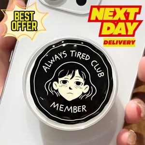 May include: A black and white phone grip with the text "ALWAYS TIRED CLUB MEMBER" and a cartoon illustration of a tired person. The grip is round and attached to a white phone.