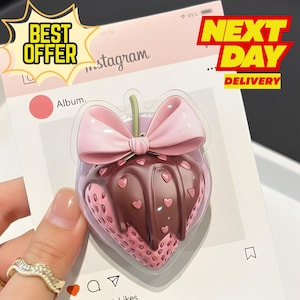 Può includere: Un supporto per telefono a forma di cuore a tema fragola con fiocco rosa e copertura al cioccolato. Il supporto è su un supporto rosa a tema Instagram. Le parole "BEST OFFER" e "NEXT DAY DELIVERY" sono visibili.