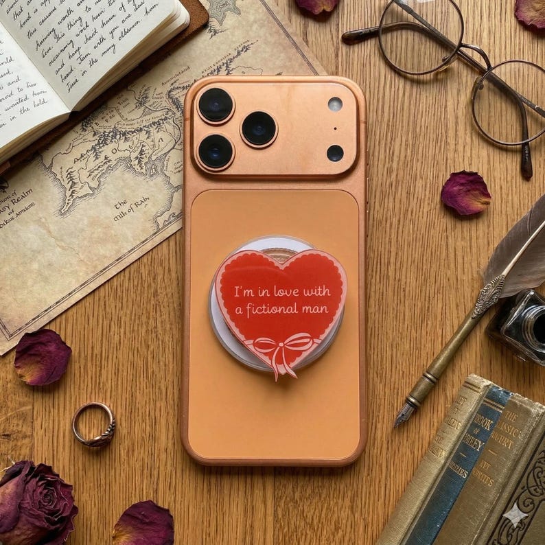 Puede incluir: Una funda de tel&eacute;fono color melocot&oacute;n con un agarre para tel&eacute;fono en forma de coraz&oacute;n rojo que dice "I'm in love with a fictional man". La imagen est&aacute; sobre una superficie de madera con libros, gafas y p&eacute;talos de rosa secos.