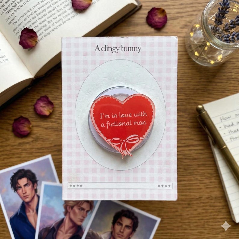Puede incluir: Un soporte para tel&eacute;fono en forma de coraz&oacute;n rojo con el texto "I'm in love with a fictional man" y un dise&ntilde;o de lazo. El soporte est&aacute; sobre un fondo a cuadros rosa y blanco, con las palabras "A clingy bunny" en la parte superior.