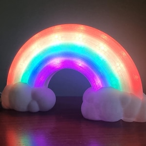 May include: A rainbow-shaped night light emitting a soft glow. The rainbow has bands of red, orange, yellow, green, blue, and purple. White cloud-shaped bases support the rainbow. The light is on, illuminating the room.