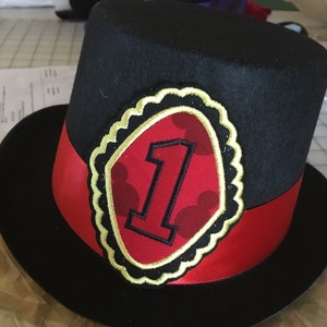 May include: Black top hat with a red and gold trim. The hat has a red and gold patch with a number one and Mickey Mouse ears on it.