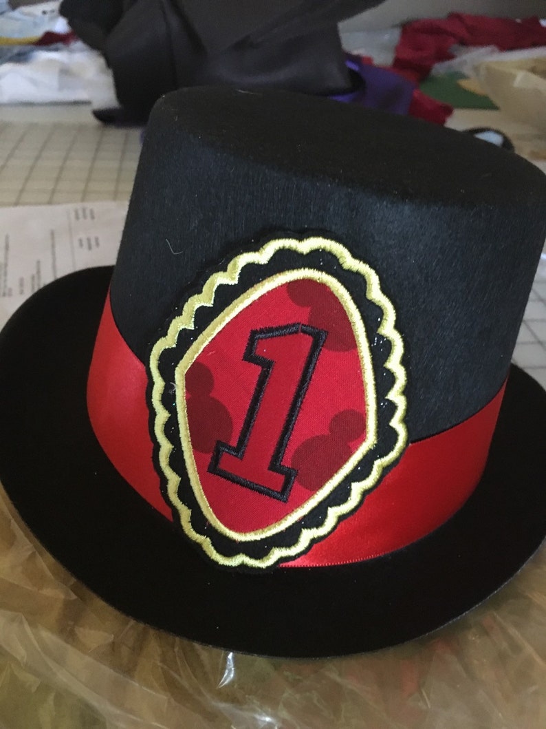 May include: Black top hat with a red and gold trim. The hat has a red and gold patch with a number one and Mickey Mouse ears on it.