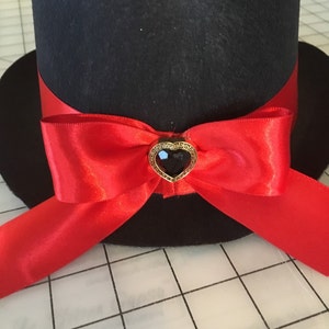May include: A black top hat with a red satin ribbon tied in a bow. The bow has a gold heart-shaped brooch with a black gem in the center.