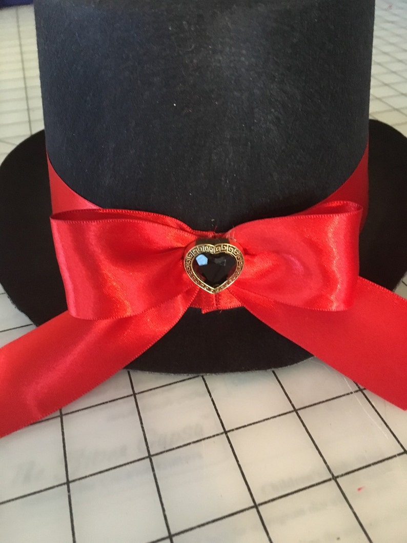 May include: A black top hat with a red satin ribbon tied in a bow. The bow has a gold heart-shaped brooch with a black gem in the center.