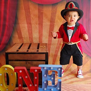 May include: A baby boy dressed as a ringmaster in a red and black costume with a top hat and bow tie. The baby is standing in front of a red and gold circus backdrop with the word "ONE" spelled out in large, colorful letters.