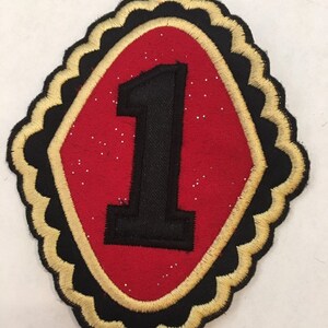 May include: A red felt patch with a black embroidered number "1" inside a gold and black scalloped border.