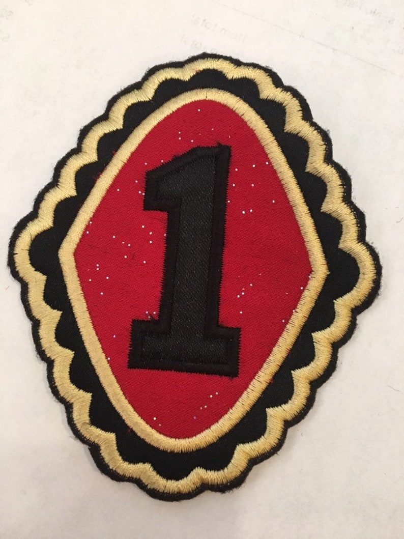 May include: A red felt patch with a black embroidered number "1" inside a gold and black scalloped border.