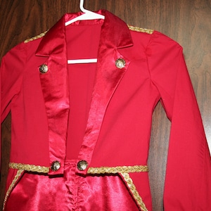 Puede incluir: Una chaqueta roja con ribetes y botones dorados. La chaqueta tiene un cuello de sat&eacute;n y cuello en V.