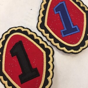May include: Two red felt patches with gold trim and black or blue felt number ones. The patches have a scalloped edge.