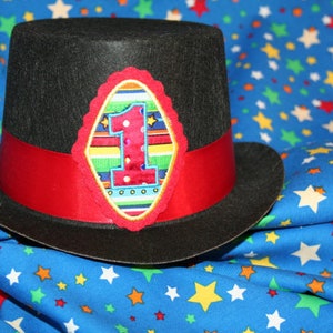 May include: A black top hat with a red band and a colorful embroidered patch featuring the number "1" on a blue background. The hat is sitting on a blue fabric with white stars.