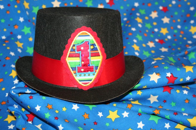 May include: A black top hat with a red band and a colorful embroidered patch featuring the number "1" on a blue background. The hat is sitting on a blue fabric with white stars.