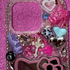 K&ouml;nnte beinhalten: Eine transparente Handyh&uuml;lle, verziert mit verschiedenen Charms. Zu den Charms geh&ouml;ren ein pinkfarbener High Heel, ein Kreuz, ein Hello Kitty-Design, Herzen und eine 8-Ball. Die H&uuml;lle ist mit Strasssteinen und anderen dekorativen Elementen verziert.