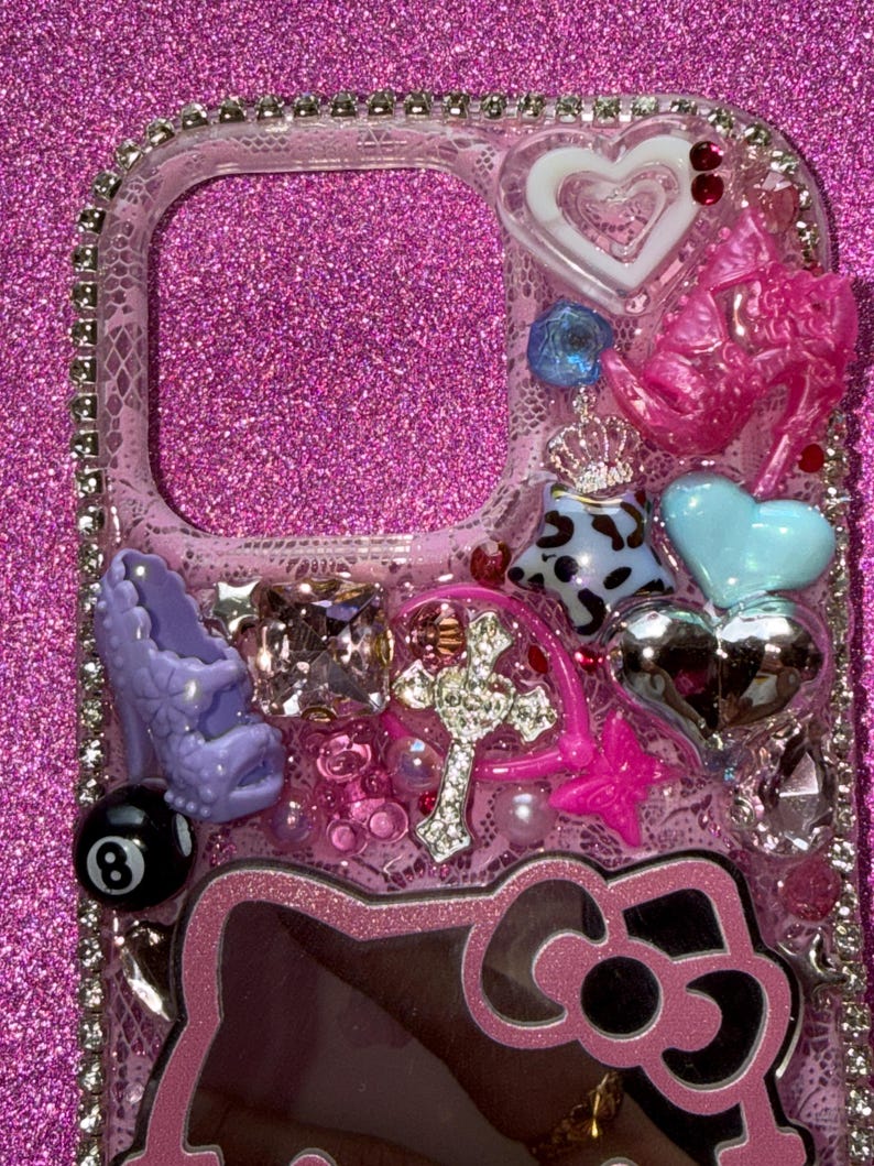 K&ouml;nnte beinhalten: Eine transparente Handyh&uuml;lle, verziert mit verschiedenen Charms. Zu den Charms geh&ouml;ren ein pinkfarbener High Heel, ein Kreuz, ein Hello Kitty-Design, Herzen und eine 8-Ball. Die H&uuml;lle ist mit Strasssteinen und anderen dekorativen Elementen verziert.