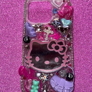 K&ouml;nnte beinhalten: Eine dekorative Handyh&uuml;lle mit pinkfarbenem Glitzerhintergrund. Die H&uuml;lle zeigt ein Hello Kitty-Design, ein Kreuz, einen High-Heel-Schuh, eine Handtasche, einen Teddyb&auml;ren und andere Verzierungen. Die H&uuml;lle ist mit Strasssteinen besetzt.