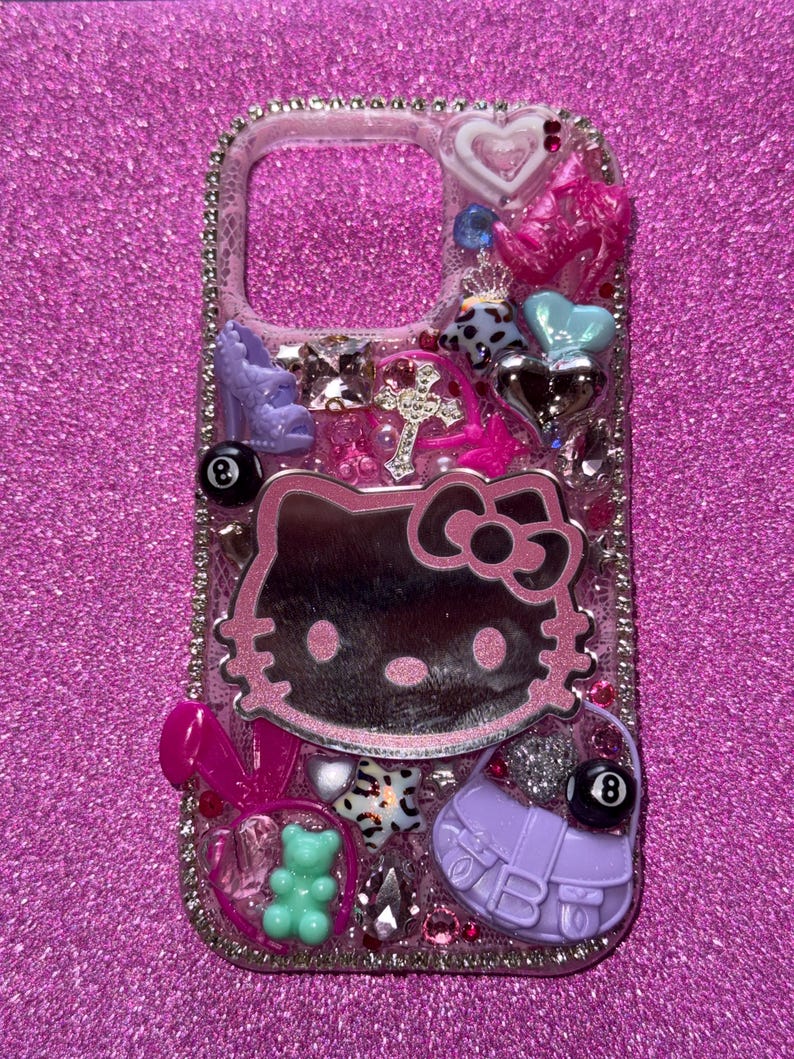 K&ouml;nnte beinhalten: Eine dekorative Handyh&uuml;lle mit pinkfarbenem Glitzerhintergrund. Die H&uuml;lle zeigt ein Hello Kitty-Design, ein Kreuz, einen High-Heel-Schuh, eine Handtasche, einen Teddyb&auml;ren und andere Verzierungen. Die H&uuml;lle ist mit Strasssteinen besetzt.