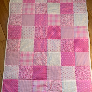 May include: A patchwork quilt featuring a grid of pink and white squares. The squares display various patterns, including floral prints, stripes, and polka dots. The quilt is bordered with a white fabric.