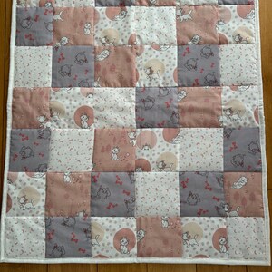 Op de afbeelding: Een patchwork quilt met een herhalend patroon van roze, grijze en witte vierkanten. Elke vierkant toont een kattenillustratie, sommige bevatten ook een cirkelvormig ontwerp. De quilt heeft een witte rand. Afmetingen ca. 70 cm x 70 cm.