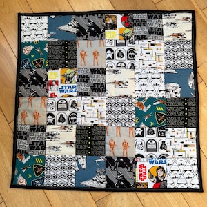 May include: A patchwork quilt with Star Wars themed fabric squares. The squares show characters, spaceships, and logos from the Star Wars franchise. The quilt has a black border and is set on a wooden surface.