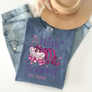 Può includere: Una maglietta blu con il Gatto del Cheshire di Alice nel Paese delle Meraviglie e la scritta "You May Have Noticed, I'm Not All There." L'immagine include jeans chiari, un cappello beige e una collana d'oro.
