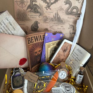 May include: A themed gift box containing a map, a "Beware" book, a notebook, a feather, a bottle of ink, chocolate coins, and other themed items. The box is filled with gold-coloured shredded paper.