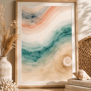 Könnte beinhalten: Aquarelldruck einer Strandlandschaft mit geschichteten Wellen in Türkis, Pfirsich und Beige. Das Kunstwerk ist in einem hellen Holzrahmen gerahmt. Eine Muschel und Bücher mit den Titeln "The Kinfolk Home", "Coastal Living" und "This is Home" befinden sich im Vordergrund.