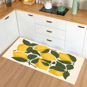 May include: A rectangular kitchen mat with a lemon design. The mat has a cream background with illustrations of yellow lemons and green leaves. The mat is placed in front of white kitchen cabinets and a wooden countertop.