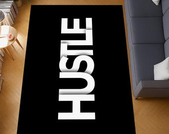 Puede incluir: Una alfombra rectangular negra con la palabra "HUSTLE" en letras blancas grandes con efecto 3D. La alfombra está colocada sobre un suelo de madera, con un sofá gris y una pequeña mesa en el fondo.