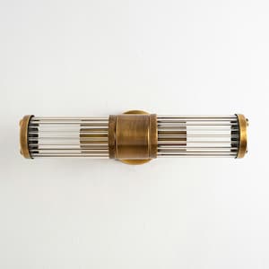 May include: A horizontal light fitting with an antique brass finish. The design incorporates a central brass plate and rounded brass end caps. Clear glass rods are visible within the fixture. The overall length is approximately 30 cm, creating a decorative lighting element.