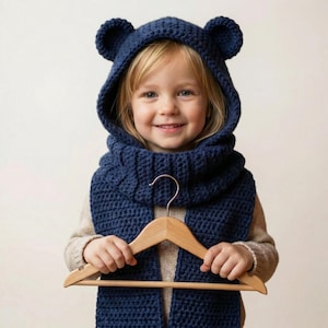 Pode incluir: Uma criança usando um conjunto de gorro e cachecol de urso de crochê azul marinho. O gorro tem orelhas de urso. A criança segura um cabide de madeira. O conjunto é feito de fio grosso e texturizado.